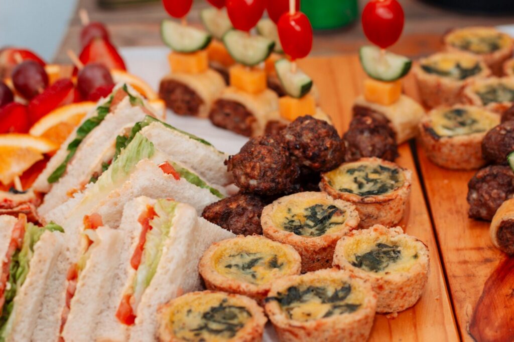 Assorted finger foods including sandwiches, quiches, and kebabs on a wooden platter, ready to serve.